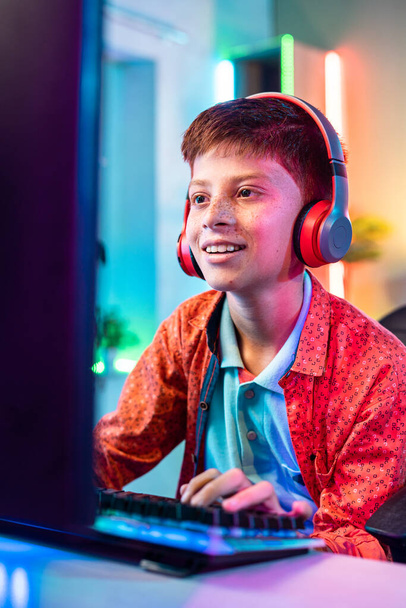 Vertikale Aufnahme, fröhlich lächelndes Teenager-Kind, das Live-Videospiel am Computer spielt, indem es zu Hause mit drahtlosem Headset spricht - Konzept der Unterhaltung, Technologiesucht und Cyberspace. - Foto, Bild