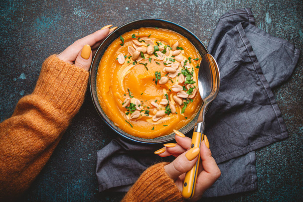 Female hands in yellow knitted sweater holding a bowl with pumpkin cream soup on dark stone background with spoon decorated with peanuts, top view. Autumn cozy dinner concept . - Photo, Image