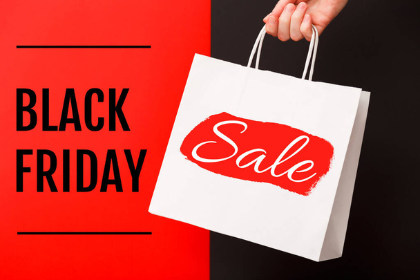 Female hand holding white shopping paper bag with Black Friday Sale text on red and black background. Sale, discount, shopping concept - Fotó, kép