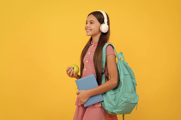 dítě ve sluchátkách se sešitem a jablkem k obědu. 1. září zdravé dětství. dítě se školní taškou. hudba. usmívající se dívka nosit batoh. Zpátky do školy. Den znalostí. koncepce vzdělávání. - Fotografie, Obrázek