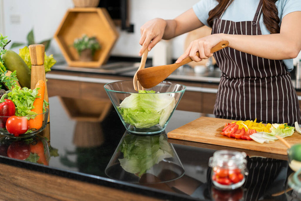 Nahaufnahme einer Asiatin mit Schürze, die den vegetarischen Salat mit Holzspachteln wirft. Zubereitung eines gesunden Salats mit frischem Gemüse wie Karotten, Tomatenkohl und grüner Eiche. - Foto, Bild