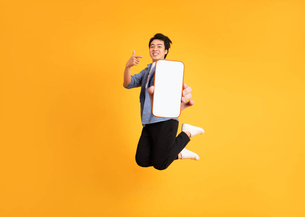 image of jumping up asian man holding smartphone in hand, isolated on yellow background - Фото, зображення