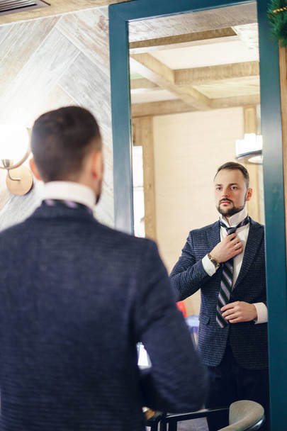 Un jeune homme avec une petite barbe ajuste sa cravate en regardant dans un grand miroir. Jeune homme élégamment habillé. - Photo, image
