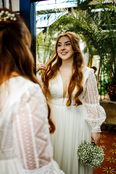 Šťastná nevěsta s rudými vlasy v elegantních šatech při pohledu do zrcadla. - Fotografie, Obrázek