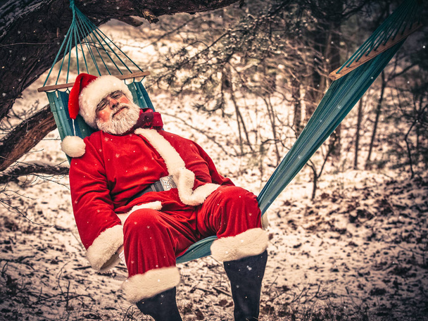 Viejo Santa Claus cansado en un traje tradicional rojo duerme en una hamaca en un acogedor bosque oscuro de invierno. - Foto, Imagen