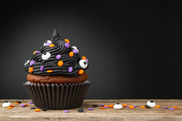 カップケーキだ。ハロウィンの黒カップケーキ。ハロウィンパーティーのデザート。チョコレートマフィンは、色のスプリンクル、霜降りとアイシングで飾られました。木製のテーブルの上にカップケーキ。暗い背景とコピースペース.  - 写真・画像