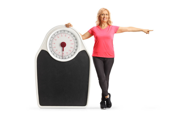 Full length portrait of a mature woman in sportswear posing with a big measuring and pointing to the side isolated on white background - Foto, Imagem