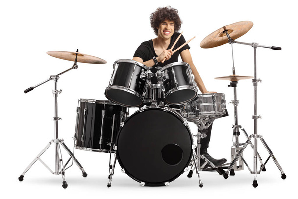 Smiling young male drummer holding drumsticks and sitting with a drum set isolated on white background - Foto, Imagem