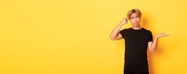Disappointed asian guy with blond hair, raising hand up puzzled and scolding someone acting stupid, standing yellow background. - Foto, afbeelding