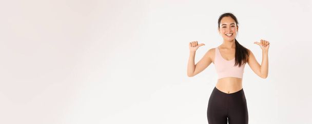 Sport, wellbeing and active lifestyle concept. Proud and happy smiling asian female fitness instructor, sportswoman pointing at herself, gaining workout goal, become gym member, white background. - Photo, Image