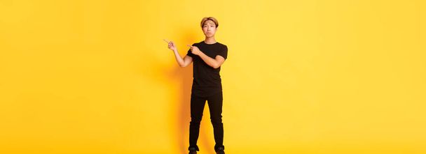 Full-length of sad and miserable asian guy in black clothes, pouting, pointing fingers upper right corner, standing yellow background. - Fotografie, Obrázek