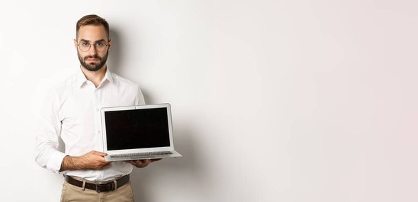 Confident manager demonstrating presentation on screen, showing laptop display and looking serious, standing over white background. - Photo, image