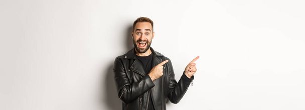 Fiducioso macho uomo in giacca di pelle nera, puntando il dito sinistro all'offerta promozionale, mostrando logo, sfondo bianco. - Foto, immagini