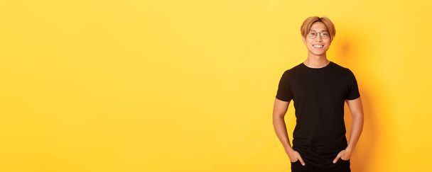 przystojny Azji blondynka facet w okulary stojąc w czarny ubrania i uśmiechając zadowolony żółty tło. - Zdjęcie, obraz