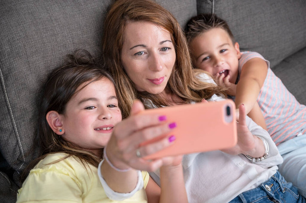 Happy mother with her kids are making a selfie or video call to father or relatives in a sofa. Concept of technology, new generation, family, connection, parenthood. High quality 4k footage - 写真・画像