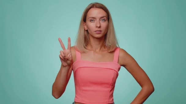Blonde slim thin slender woman showing victory sign, hoping for success and win, doing peace gesture, smiling with kind optimistic expression. Young adult girl isolated on blue studio wall background - Footage, Video