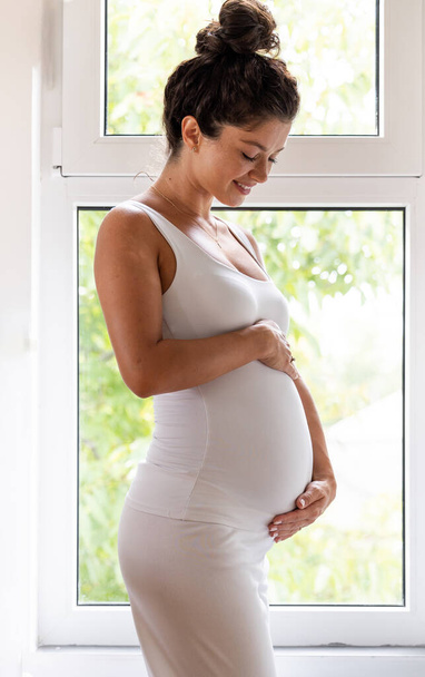 Beautiful pregnant woman in white holding hands on belly in front of window. Maternity period and baby love concept - Foto, imagen