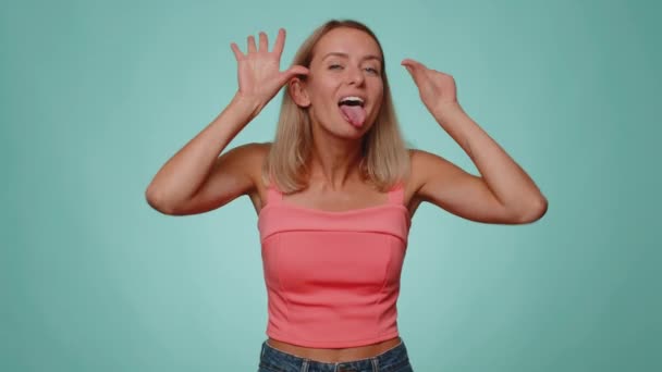 Cheerful funny slim thin slender woman showing tongue making faces at camera, fooling around, joking, aping with silly face, teasing. Young pretty adult girl isolated alone on blue studio background - Footage, Video