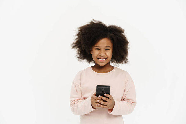 Black preteen girl with curly hair smiling and using cellphone isolated over white background - Photo, image