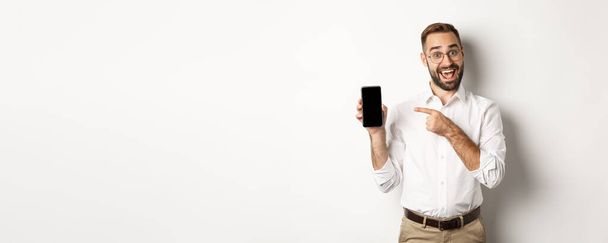 Ενθουσιασμένος όμορφος άντρας που δείχνει κινητό τηλέφωνο, δείχνει με το δάχτυλο την οθόνη και χαμογελά, στέκεται πάνω σε λευκό φόντο. - Φωτογραφία, εικόνα