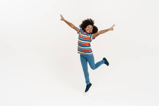 Black preteen girl smiling and showing peace gesture while jumping isolated over white background - Foto, imagen