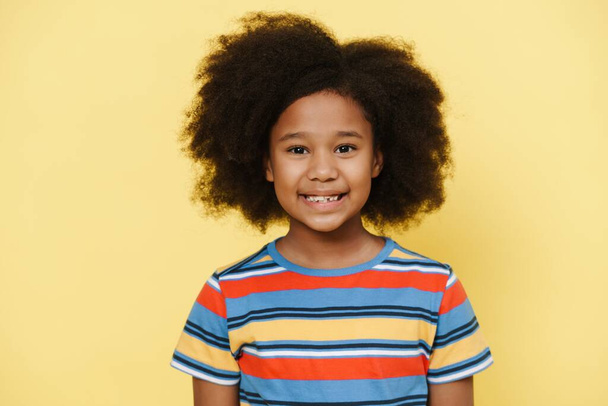 Black preteen girl with curly hair smiling and looking at camera isolated over yellow background - Фото, изображение
