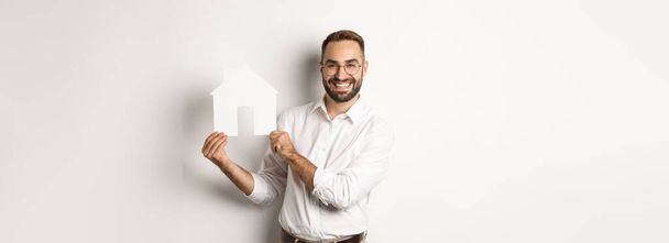 Real estate. Handsome man showing house model and smiling, broker showing apartments, standing over white background. - 写真・画像