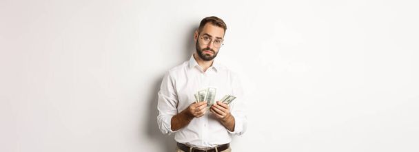 Handsome businessman counting money and looking at camera, standing serious against white background. - Photo, Image