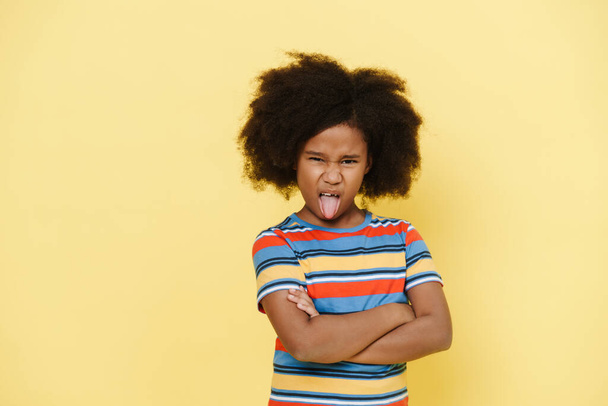 Black preteen girl frowning while showing her tongue at camera isolated over yellow background - Fotoğraf, Görsel