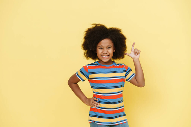 Black preteen girl with curly hair smiling and pointing finger upward isolated over yellow background - Foto, immagini