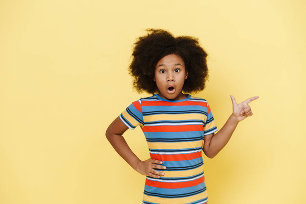 Black preteen girl expressing surprise and pointing finger aside isolated over yellow background - Foto, Imagem
