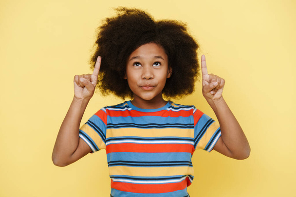 Black preteen girl with curly hair pointing fingers upward isolated over yellow background - Valokuva, kuva