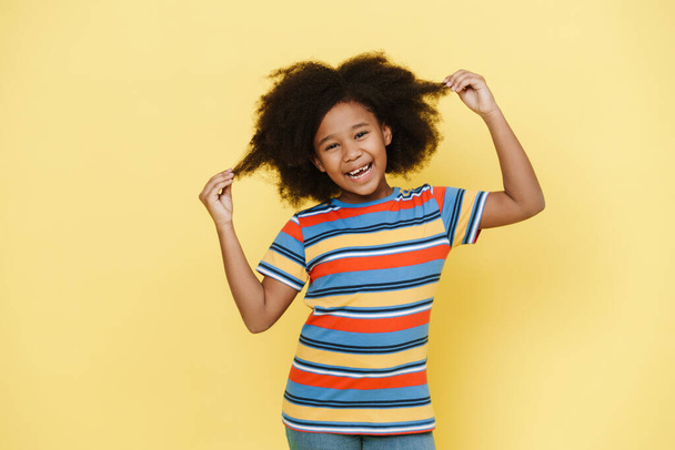 Black preteen girl laughing while holding her hair isolated over yellow background - Foto, Bild