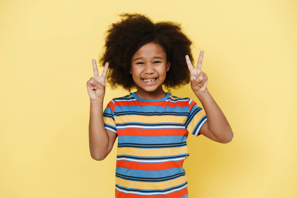 Black preteen girl with curly hair smiling and showing peace gesture isolated over yellow background - Zdjęcie, obraz