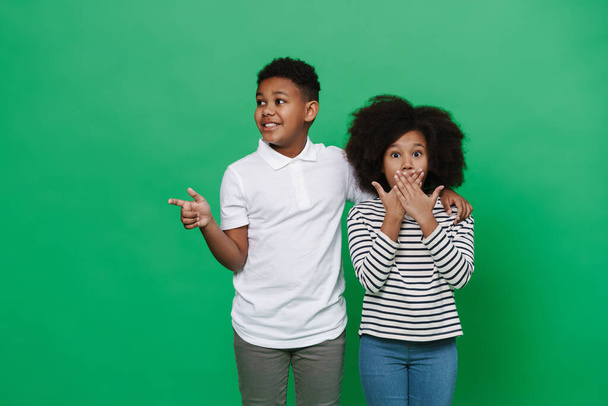 Black boy and girl expressing surprise while pointing finger aside isolated over green background - Foto, Imagem