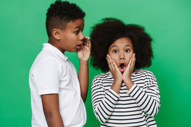 Black boy whispering secret to his shocked friend isolated over green background - Fotoğraf, Görsel