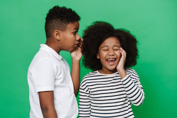 Black boy whispering secret to his laughing friend isolated over green background - Фото, изображение