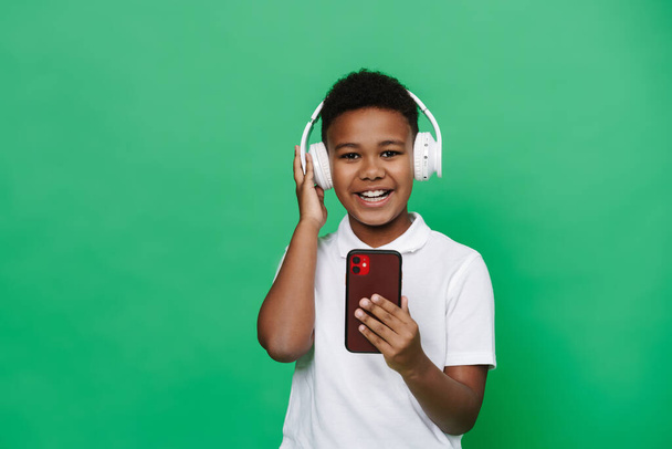 Black preteen boy listening music with headphones and cellphone isolated over white background - Foto, Imagem