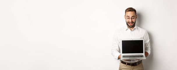 Opgewonden zakenman toont iets op laptop scherm, staande blij over witte achtergrond. - Foto, afbeelding