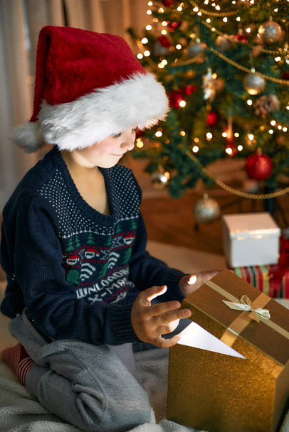 Szczęśliwy chłopiec w kapeluszu Mikołaja otwiera pudełko z prezentem świątecznym obok choinki w girlandach. Szczęśliwego Nowego Roku i świąt Bożego Narodzenia.  - Zdjęcie, obraz