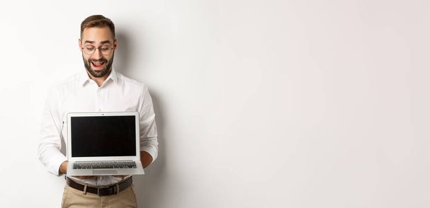 Emocionado hombre de negocios mostrando algo en la pantalla del ordenador portátil, de pie feliz sobre el fondo blanco. - Foto, imagen