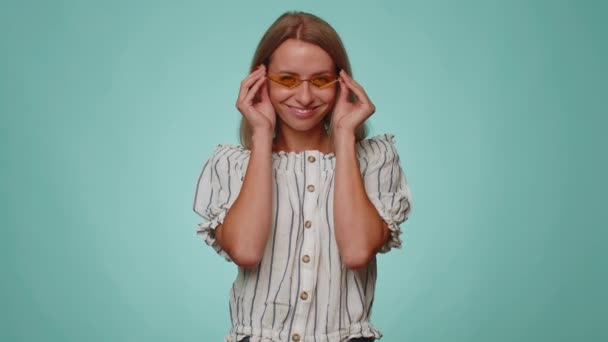 Verspielt glücklich blonde erwachsene Frau 20 Jahre alt mit gelber Sonnenbrille blinkenden Augen in die Kamera mit zahmem Lächeln, zwinkert und flirtet Ausdruck von Optimismus. Junge hübsche Mädchen auf blauem Hintergrund - Filmmaterial, Video