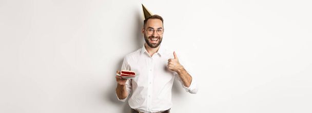 Svátky a oslavy. Spokojenost muž těší b-day party, držení narozeninového dortu a ukazující palec nahoru v souhlasu, doporučující něco, bílé pozadí. - Fotografie, Obrázek