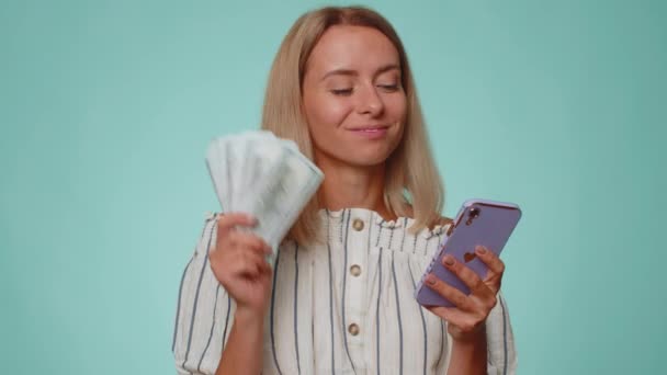 Happy tourist woman looking smartphone display sincerely rejoicing win, receiving money dollar cash banknotes, success lottery luck. Young pretty blonde girl isolated alone on blue studio background - Footage, Video