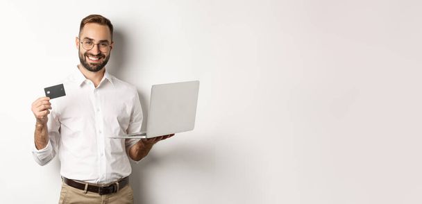 Online shopping. Handsome man showing credit card and using laptop to order in internet, standing over white background. - Foto, imagen