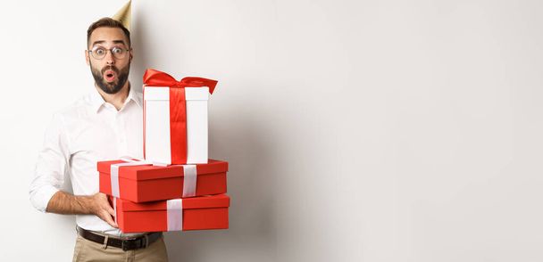 Γιορτές και γιορτή. Ευτυχισμένος άνθρωπος λαμβάνει δώρα για τα γενέθλια, κρατώντας δώρα και αναζητούν ενθουσιασμένοι, στέκεται πάνω από το λευκό φόντο. - Φωτογραφία, εικόνα