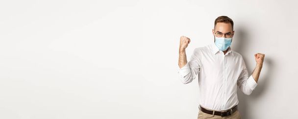 Covid-19, social distancing and quarantine concept. Confident business man in medical mask fist pump, rejoicing of winning, triumphing while standing over white background. - Foto, imagen