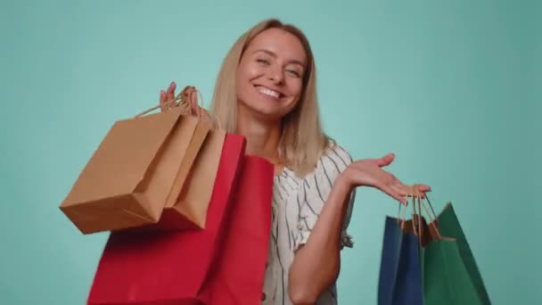 Χαρούμενη τουρίστρια που δείχνει τσάντες για ψώνια, διαφημιστικές εκπτώσεις, χαμογελώντας δείχνοντας έκπληκτος με χαμηλές τιμές, ψώνια στις διακοπές της Μαύρης Παρασκευής. Νεαρή ξανθιά λεπτή λεπτή λεπτή κοπέλα σε μπλε φόντο - Πλάνα, βίντεο