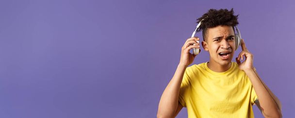 Cant hear you, repeat please. Portrait of young bothered guy interrupted of listening music, take-off headphones to answer person question, squinting look confused, purple background. - Photo, Image