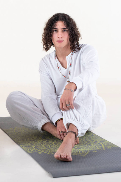 Egy fiatal férfi ül a szőnyegen meditálni, miközben a kamerát nézi fehér háttér felett. Stúdió felvétel. - Fotó, kép